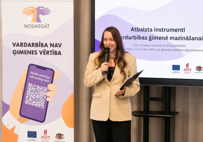 Persona gaišā žaketē stāv pie mikrofona un planšetes konferenču telpā, uzstājoties pasākumā par vardarbības ģimenē mazināšanu. Blakus redzams informatīvs stends ar uzrakstu “Vardarbība nav ģimenes vērtība” un QR kodu, bet fonā ekrānā attēlota prezentācija par atbalsta instrumentiem vardarbības ģimenē mazināšanai. Redzami Eiropas Savienības un Latvijas institūciju logotipi.