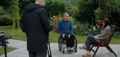Vīrietis ratiņkrēslā, sieviete, intervētāja, dārza krēslā un operators pie TV kameras mājas dārza vidē. 