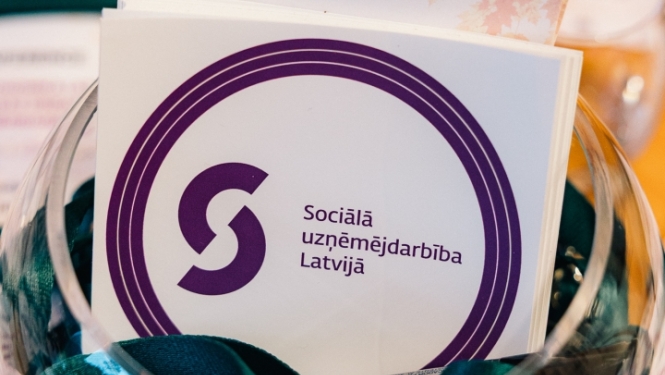 Fotogrāfijā redzams caurspīdīgs trauks ar tumši zaļiem auduma maisiņiem un informatīvajiem materiāliem par sociālo uzņēmējdarbību Latvijā. Priekšplānā redzams buklets ar uzrakstu “Sociālā uzņēmējdarbība Latvijā”, bet fonā – citi informatīvie materiāli.