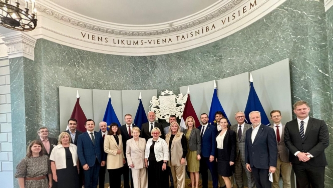 Grupas foto reprezentatīvā telpā ar Latvijas un Eiropas Savienības karogiem fonā. Aptuveni 20 cilvēki stāv puslokā zem Latvijas valsts ģerboņa. Virs grupas uz sienas redzams uzraksts: “Viens likums – viena taisnība visiem”. Telpai raksturīgas zaļganas marmora sienas un klasiska interjera elementi.