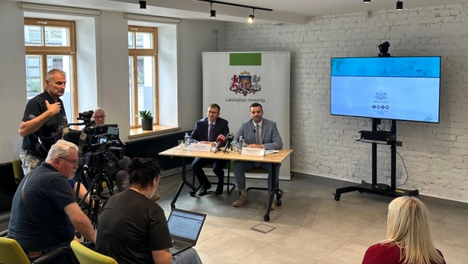 Preses konference gaišā telpā ar balti krāsotu ķieģeļu sienu. Priekšplānā sēž žurnālisti un operatori ar kamerām un klēpjdatoriem. Telpas aizmugurē pie galda sēž divi vīrieši, bet aiz viņiem redzams Latvijas Labklājības ministrijas logotips. Labajā pusē uz ekrāna redzama prezentācija, bet pie griestiem izvietoti melni sliežu prožektori.