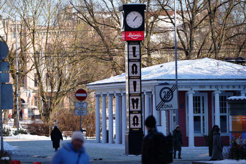 Foto redzams Rīgas Laimes pulkstenis, kas ir saldākā satikšanās vieta Rīgā. Redzama ikdienas aina, kur cilvēki staigājas gar laimas pulksteni.