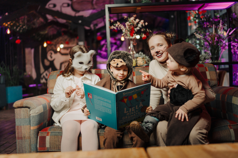 pasaku festivāla vizuālais attēls. Bērni dzivnieku kostimos sēž un lasa grāmatu