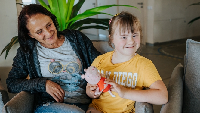 Mamma ar bērnu sēž istabā ar baltām sienām. Fonā liels telpaugs