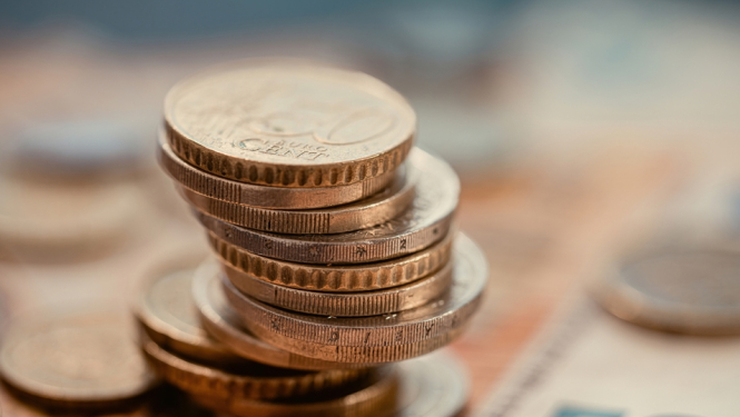 Kaudzē sakrautas eiro monētas, kas novietotas uz izpludināta 50 eiro banknotes fona.