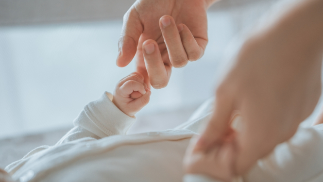 Pieaugušā roka maigi tur jaundzimušā roku, simbolizējot rūpes un drošību.