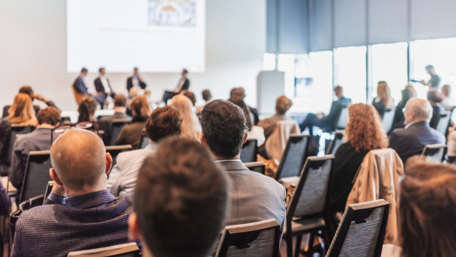 Konference vai seminārs ar auditoriju, kurā cilvēki sēž un klausās paneļdiskusiju priekšā uz skatuves.