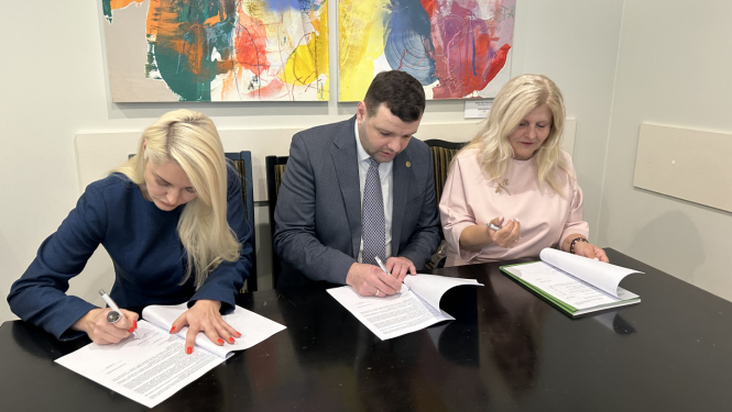 Divas sievietes un viens vīrietis (Labklājības ministrs) sēž pie tumša galda un paraksta dokumentus, koncentrējoties uz papīriem; aiz viņiem pie sienas ir divi krāsaini abstraktās mākslas darbi.