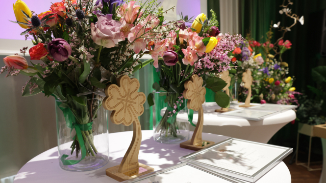 Ilustratīvs attēls - Krāsainu ziedu pušķi stikla vāzēs uz galda, blakus novietoti ierāmēti atzinības raksti un koka dekoratīvi elementi āboliņa formā pasākuma telpā.