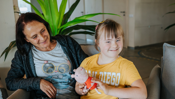 Mamma ar bērnu sēž istabā ar baltām sienām. Fonā liels telpaugs