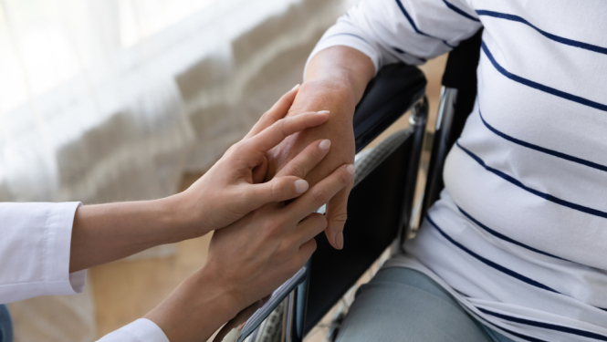 Cilvēks ratiņkrēslā, kura roku atbalstoši tur cita persona, izrādot līdzjūtību un rūpes.