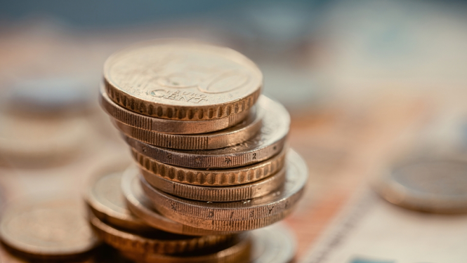 Kaudzē sakrautas eiro monētas, kas novietotas uz izpludināta 50 eiro banknotes fona.