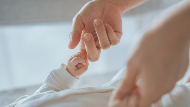 Pieaugušā roka maigi tur jaundzimušā roku, simbolizējot rūpes un drošību.