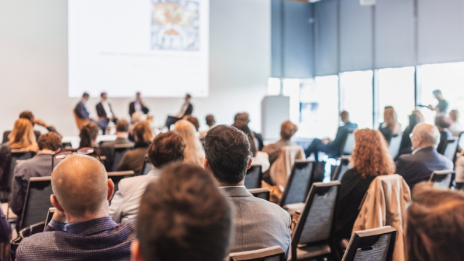 Konference vai seminārs ar auditoriju, kurā cilvēki sēž un klausās paneļdiskusiju priekšā uz skatuves.