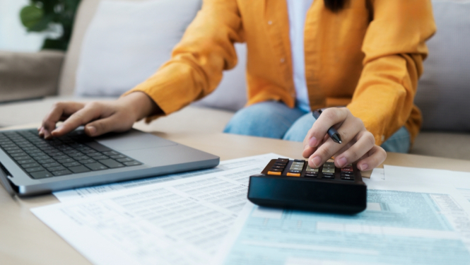 Sievietes siluets, sēž pie datora, otrā rokā kalkulators un rēķina jaunās pabalsta summas