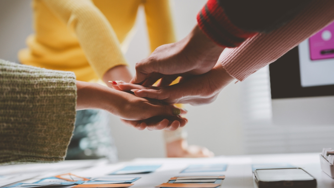 Attēlā redzamas vairākas cilvēku rokas, kas saliktas kopā virs galda kā komandas vienotības un sadarbības simbols. Fonā redzami papīri, krāsu paraugi un viedtālrunis, kas liecina par radošu vai darba vidi, iespējams, projekta plānošanu.