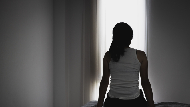 sievietes siluets no mugurpuses, sēž uz gultas un skatas pa logu, melnbalta bilde.