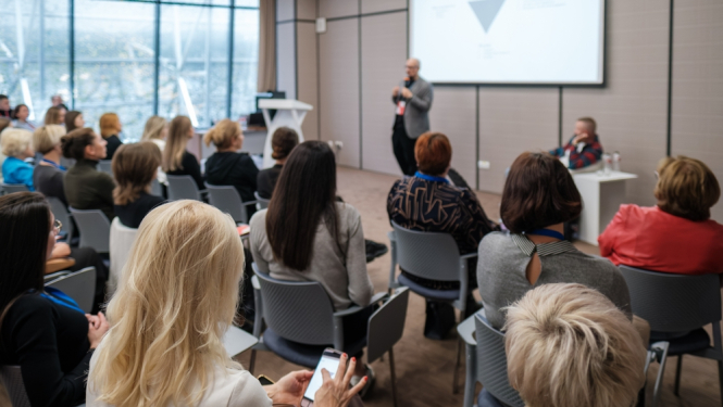 redzami daudz cilvēki no mugurpuses, kas klausās lekciju  un skatās prezentāciju kas attēlota
