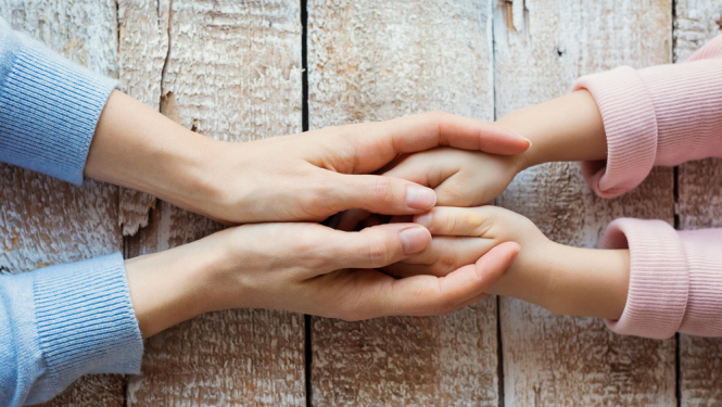 Divas pieauguša cilvēka rokas maigi tur bērna rokas, fons – balta, nolietota koka virsma.