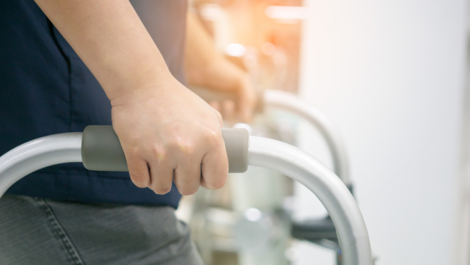 Cilvēks turas pie pārvietošanās palīglīdzekļa (staigāšanas rāmja), redzama roka uz roktura; fonā izplūdusi iekštelpu vide ar siltu apgaismojumu