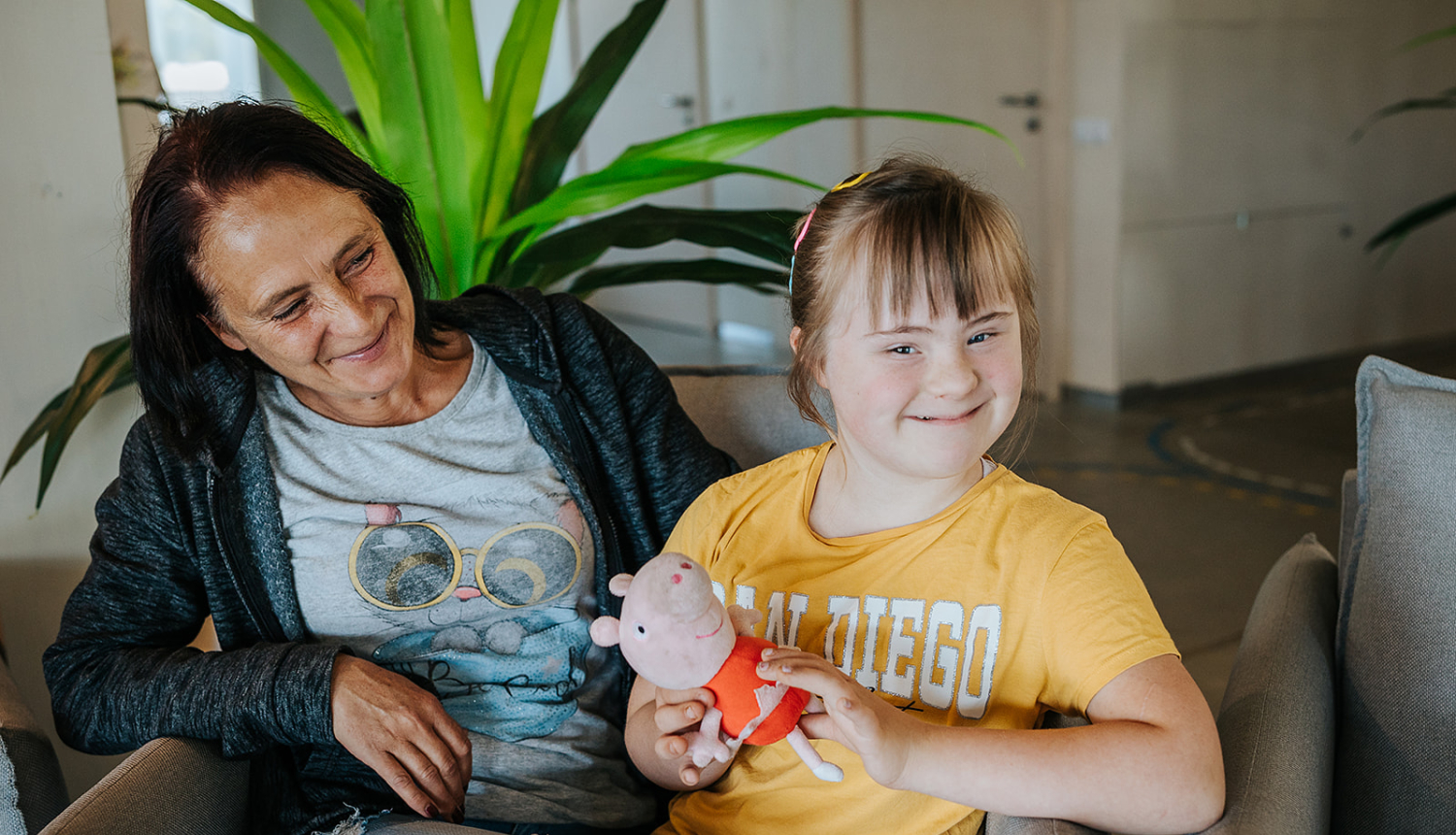 Mamma ar bērnu sēž istabā ar baltām sienām. Fonā liels telpaugs
