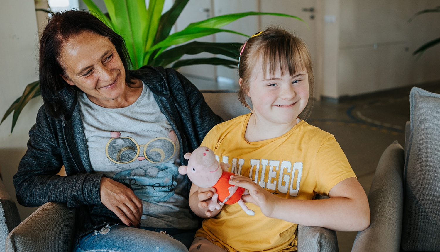 Mamma ar bērnu sēž istabā ar baltām sienām. Fonā liels telpaugs